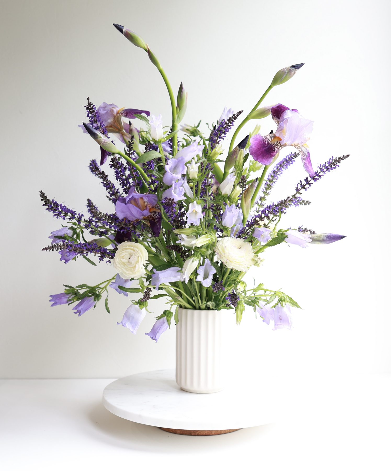 A tall purple iris, salvia, and ranunculus arrangement on a marble stand.