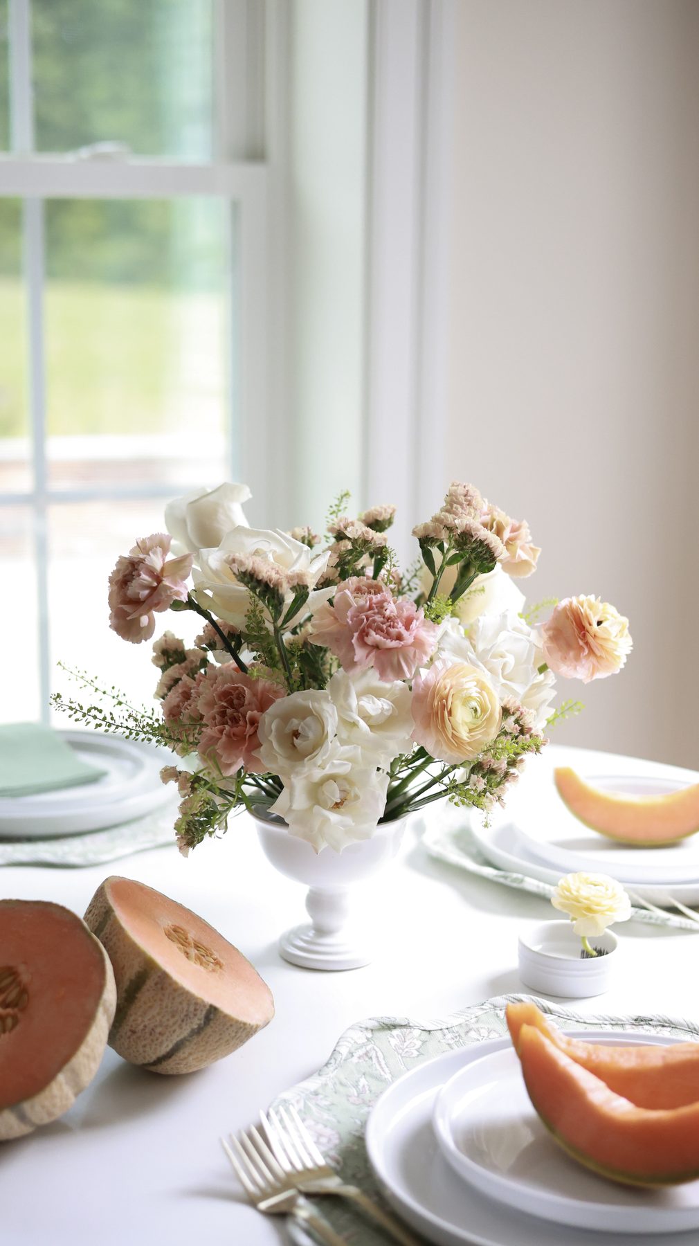 A white and blush floral arrangement on a table with fresh melon.