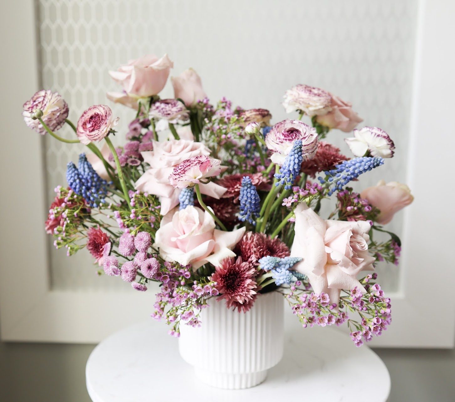 A pink rose, ranunculus, and blue muscari arrangement in a ribbed white vase.
